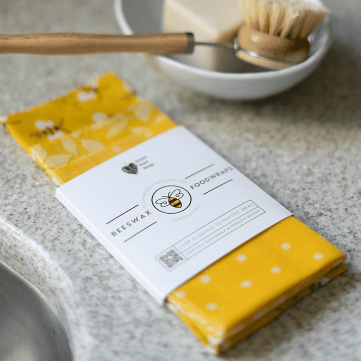 Set of three beeswax food wraps in yellow bee pattern next to a bamboo dish brush and zero waste package free dish bar soap