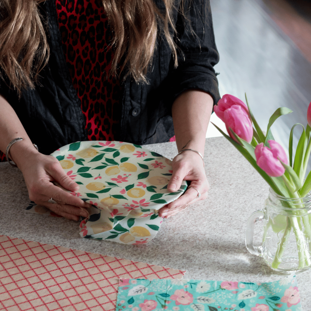 Caucasian woman wrapping bowl with Good Soul Shop beeswax wrap with lemon print - a pink and red food wrap and teal food wrap are off side